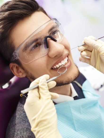 Bright young man receiving dental treatment at a clinic, wearing protective glasses, smiling, while a dentist uses dental tools for teeth cleaning or examination.