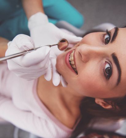 Teeth whitening procedure at a dental clinic, close-up of a woman lying down and smiling during treatment with a dental professional holding a dental mirror.